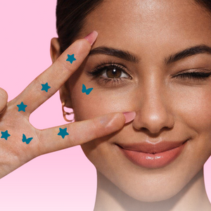 Woman making a peace sign with star-shaped stickers on her face against a pink background, promoting 'Plot Twist' 2-in-1 graphic eyeliner and stamp.